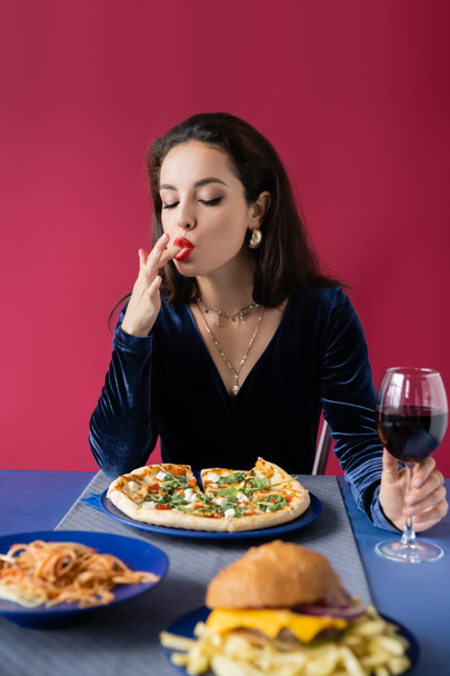 sexy woman with glass of wine licking her finger near blue table served with delicious meal isolated on red - Foto, Bild