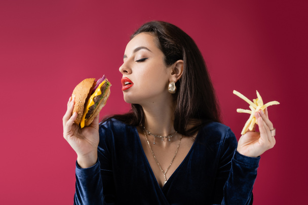 young woman in blue velour dress holding burger and french fries isolated on red - Φωτογραφία, εικόνα