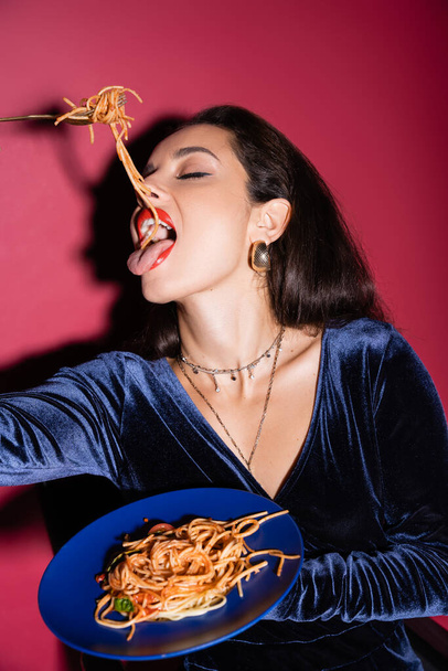 sexy woman in blue velour dress eating tasty spaghetti on red background - Фото, изображение