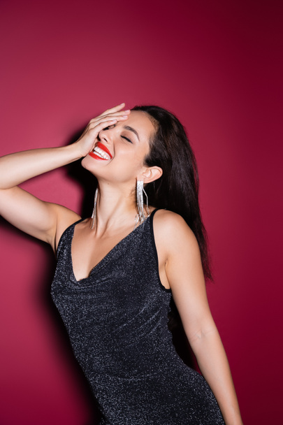 elegant woman in shiny earrings and black lurex dress obscuring face and laughing on red background - Zdjęcie, obraz