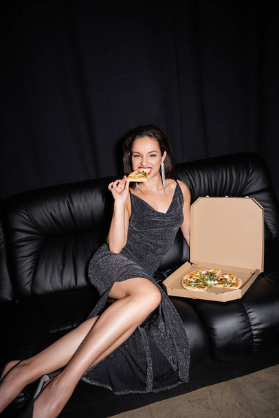 sexy woman eating delicious pizza while sitting on leather couch isolated on black - Photo, Image
