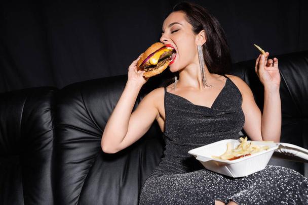 jeune femme en robe lurex manger hamburger près de récipient en plastique avec des frites isolées sur noir - Photo, image