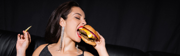 brunette femme avec les yeux fermés manger hamburger savoureux isolé sur noir, bannière - Photo, image