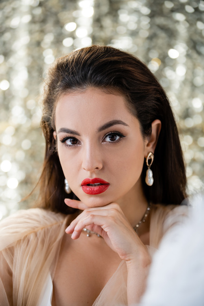 brunette woman with makeup and pearl earrings holding hand near chin and looking at camera on shiny background - Photo, Image