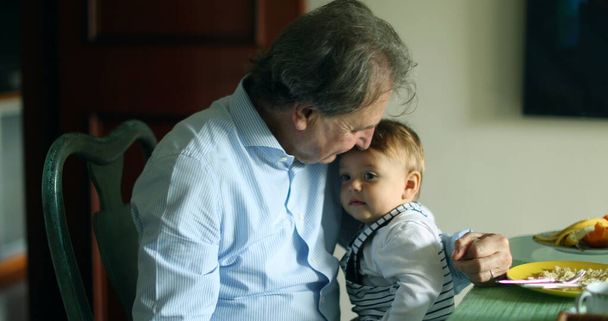 Grandfather holding grandson in arms at meal table. Casual and candid grand-parent with infant baby - Photo, image