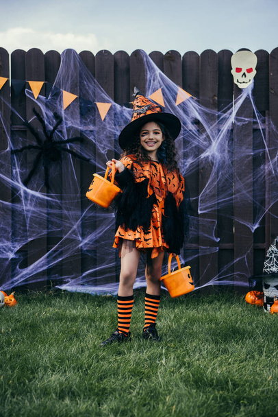 Fille souriante dans un chapeau de sorcière tenant des seaux près du décor d'Halloween sur la clôture dans la cour arrière - Photo, image