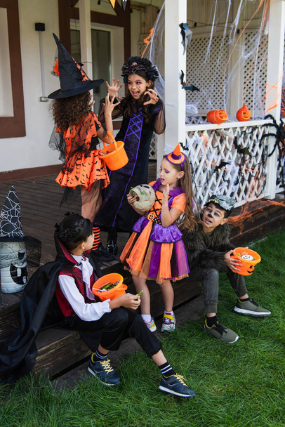 asiático chicos sentado con halloween cubos cerca niñas en bruja trajes asustando uno a otro - Foto, imagen