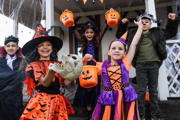 amigos multiculturales emocionados en trajes de Halloween celebración truco o tratar cubos cerca de casa - Foto, imagen