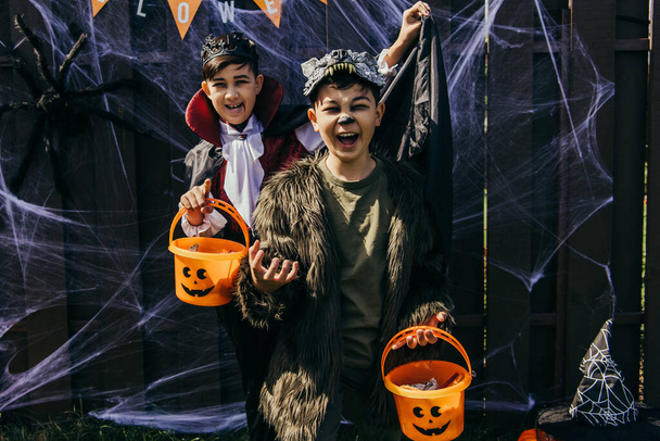 Asian kids in costumes holding halloween buckets and grimacing near fence outdoors - Foto, Imagen