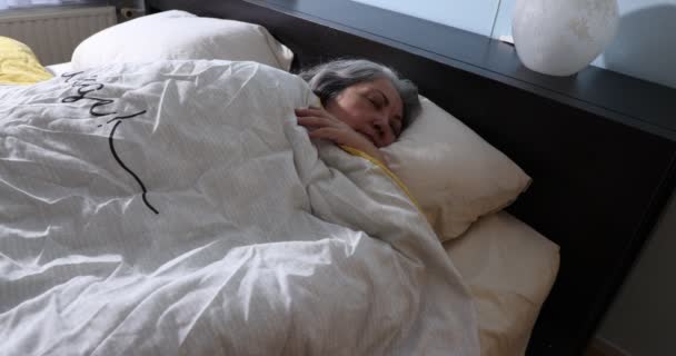 Зрелая женщина мирно спит, не желая просыпаться, чтобы продолжать мечтать, солнечный свет проходит через окно, движется медленно, длинные седые волосы, белое одеяло, абсолютное спокойствие - Кадры, видео