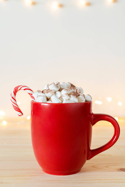 Hot chocolate in red mug with small marshmallows and candy cane on wooden table with holiday lights on wall. Cocoa in a cup, hot winter drink - Photo, Image