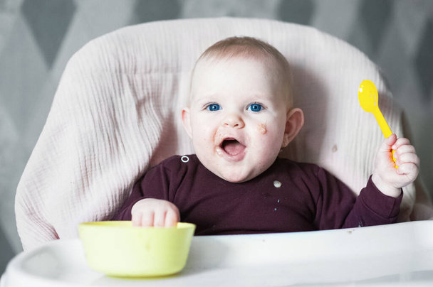 Glimlachende baby 7 maanden oud met lepel op kinderstoel - Foto, afbeelding