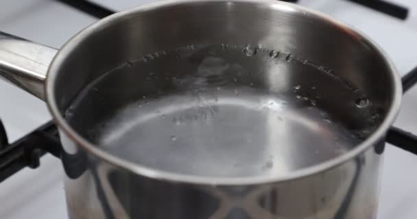 Close-up of clean transparent boiling water on a gas stove, bubbles and splashing, black grill, white stove, metal pan. Home food concept - Footage, Video