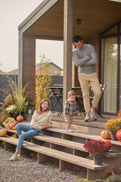 Cute little boy standing on the wooden porch steps surrounded by his father and a cheerful mother - Фото, изображение