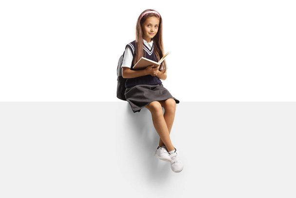Child in a school uniform sitting on a blank panel and holding a book isolated on white background - Fotó, kép