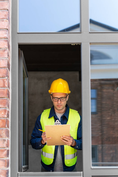 construction business and building concept - male builder in helmet and safety west with clipboard and pencil looking out window - Photo, image
