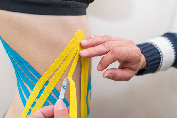 physiotherapist applying adhesive elastic therapeutic tape to the side of the abdomen of a young adult athletic woman in a doctor's office. Injury Rehabilitation and Healthcare - Photo, Image