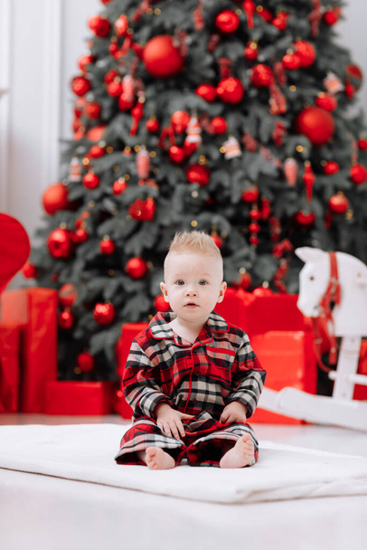 Adorable caucasian baby boy is sitting and holding gift toy Santa Claus near a Christmas tree, many festive gift boxed and red sofa at home, baby infant. Xmas and New Year celebrations concept - Foto, Bild
