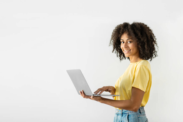 Allegro donna nera tenendo un computer portatile tra le mani e premendo il dito sulla tastiera mentre si lavora online su uno sfondo bianco parete. Spazio vuoto. Foto di alta qualità - Foto, immagini