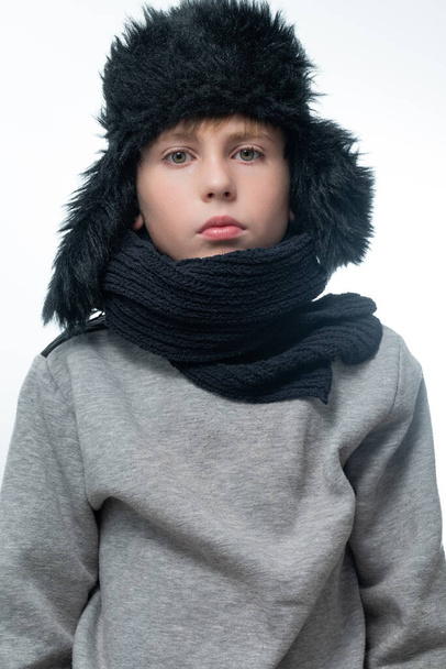 Portrait of a child in a winter hat with earflaps and a knitted scarf on a white background, black winter clothes. - Foto, immagini