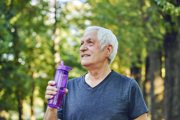 Close up image attractive mature man holds plastic reusable bottle drinking still mineral water during morning work out or stroll in summer park, caring about health, relieving thirst - 写真・画像