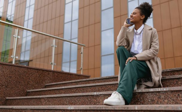 Αφροαμερικάνα νεαρή γυναίκα μιλάει στο τηλέφωνο, ενώ κάθεται στα σκαλιά με φόντο ένα επιχειρηματικό κέντρο. - Φωτογραφία, εικόνα