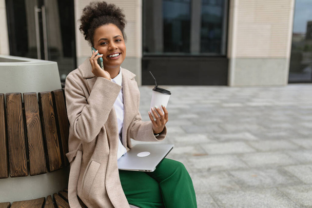 joven afroamericana mujer sentada cerca del centro de negocios durante la pausa para el almuerzo con un ordenador portátil y un vaso de café habla con una sonrisa en el teléfono. - Foto, Imagen