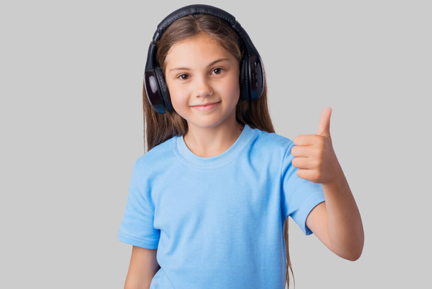 Krásná usměvavá dívka v modrém tričku poslouchat hudbu a ukazovat palce nahoru proti šedému pozadí - Fotografie, Obrázek