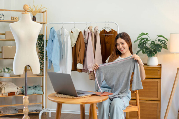 Female online seller is using laptop to broadcast live on Vlog for introduce fashions while measuring the chest size of clothes to showing for customer and selling online clothing on social media. - Fotoğraf, Görsel