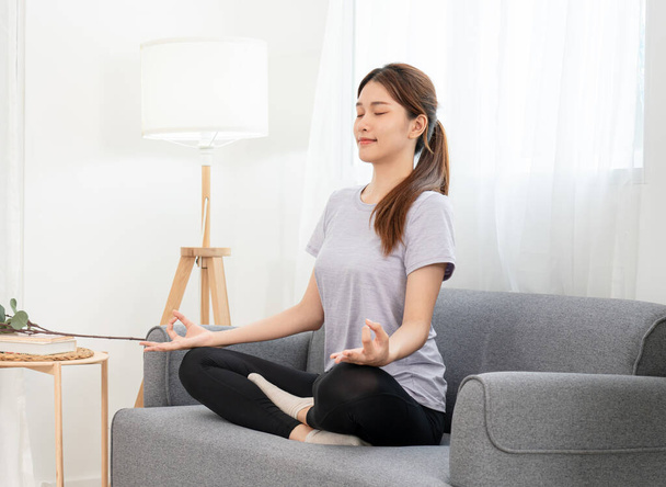 Young woman is relaxing into the lotus position for meditation on couch before workout yoga exercise in her living room at home. - 写真・画像