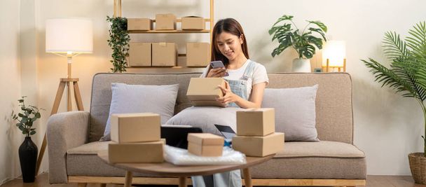 Young entrepreneur is holding cardboard boxes to taking a picture to sending customer while sitting on comfortable couch and working in living room at home office. - Фото, зображення