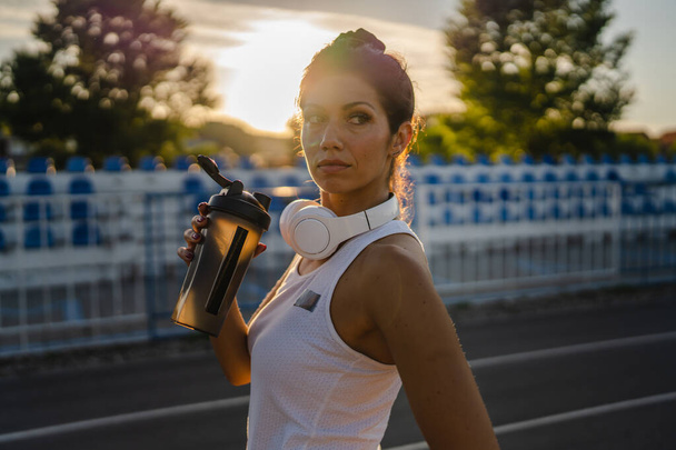 Одна жінка доросла біла тисячолітня жінка, що стоїть на стадіоні на гоночній трасі після або перед тренуванням, приймаючи гальмівну тримач додаткової пляшки з водою здоровий спосіб життя концепція копіювання простору
 - Фото, зображення