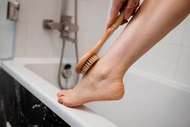Cropped photo of a young woman in white towel doing body lymphatic drainage massage with dry wooden brush with natural bristles in bathroom at home. Anti-cellulite exercises - Photo, image