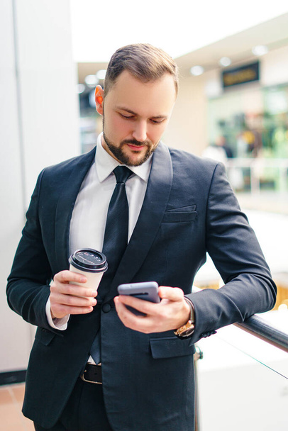 A young man with a beard in a business suit with a smartphone and a cup of coffee in the mall. Young businessman at the mall - Фото, зображення