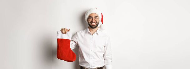 Feliz Navidad, concepto de vacaciones. Feliz hombre adulto recibir regalos en calcetín de Navidad, mirando emocionado, usando sombrero de santa, fondo blanco. - Foto, imagen