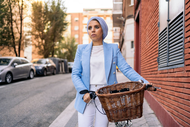 Vertical image of a muslim woman wearing a blue light suit and white pants walking with her bicycle - Foto, immagini