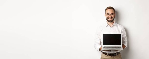 Hezký obchodník v brýlích, ukazující obrazovku notebooku a šťastný úsměv, stojící nad bílým pozadím. - Fotografie, Obrázek