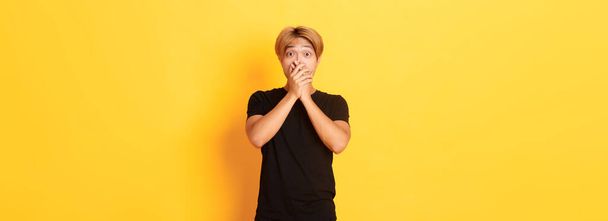 Portret zaskoczonego azjatyckiego faceta z blond fryzurą, pokrywa usta dłońmi i wygląda zdumiewająco, stojące żółte tło. - Zdjęcie, obraz