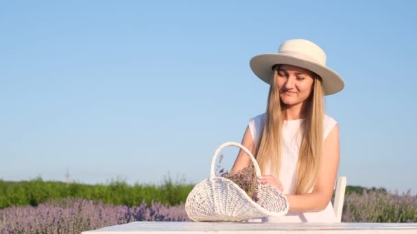 Een aantrekkelijke blonde met een witte hoed zit aan een tafel op een lavendelveld. Fotosessie in witte tinten op een lavendelveld. Provence - Video