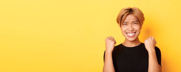 Крупним планом щасливий азіатський хлопець виграє приз, роблячи жест кулакового насоса і посміхаючись задоволені, тріумфуючи над досягненням, стоячи жовтим тлом
. - Фото, зображення