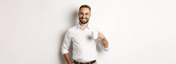 Handsome businessman drinking coffee and smiling, standing against white background. - Foto, imagen