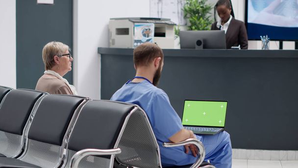 Mužská sestra analyzující zelenou obrazovku na notebooku v čekárně, sedící v nemocničním přijímacím prostoru. Prázdná šablona copyspce s maketovým pozadím a izolovaným klíčem chroma. Trojnožka. - Fotografie, Obrázek
