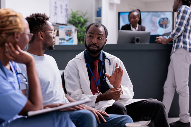 African american medical team consulting man sitting in waiting room at facility reception lobby. Doctor and assistant talking to patient about disease diagnosis and treatment, healthcare support. - Photo, Image