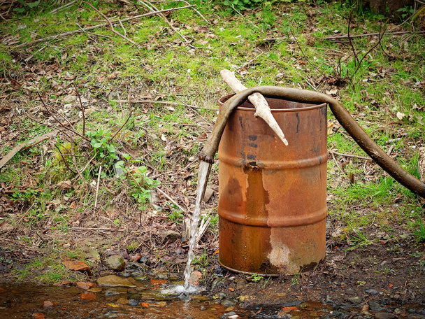 A água flui de uma mangueira deitada em um barril enferrujado na natureza - Foto, Imagem