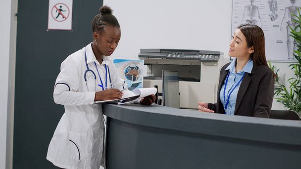 Portrait of african american doctor analyzing medical report papers with receptionist at hospital reception counter. Health specialist working at registration desk in lobby, healthcare appointments. - 写真・画像