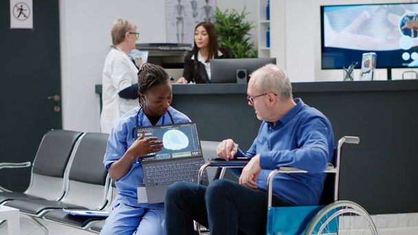 Nurse showing brain tomography to man with physical disability, doing medical consultation with patient wheelchair user. Looking at neurology scan diagnosis and neural system on laptop. - Photo, Image
