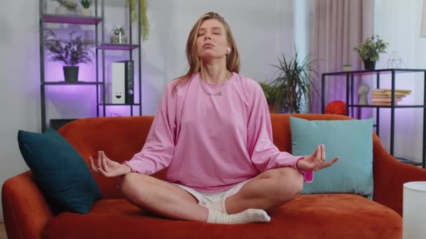 Keep calm down, relax, inner balance. Adult girl breathes deeply with mudra gesture, eyes closed meditating with concentrated thoughts, peaceful mind. Young woman sit at home in living room on couch - Materiał filmowy, wideo