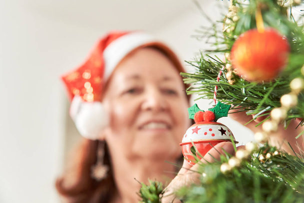 Glückliche ältere hispanische Frau lächelt, während sie zu Hause einen Weihnachtsbaum mit einer roten Nikolausmütze schmückt. Selektiver Fokus auf den Vordergrund, eine Weihnachtskugel, die sie an einem Ast hängt. - Foto, Bild