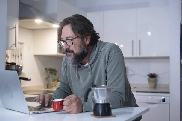 Mężczyzna w domu je śniadanie przed pracą, pije włoską kawę. Nowoczesne osoby korzystające z laptopa komputerowego w kuchni w domu prawdziwy wypoczynek stylu życia. mężczyzna osoba wewnątrz aktywność online w domu - Zdjęcie, obraz