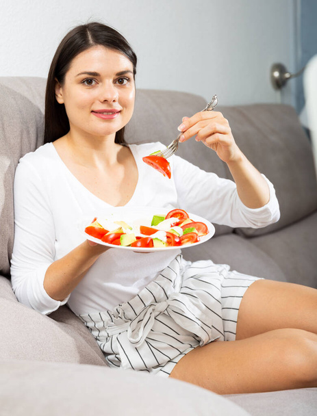 Satisfied brunette sitting on a sofa and eating salad - Photo, image